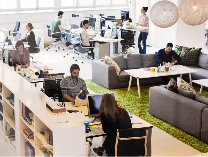 High-angle shot of a group of 8 people working with laptops, desktops, and notepads, in an open work space.